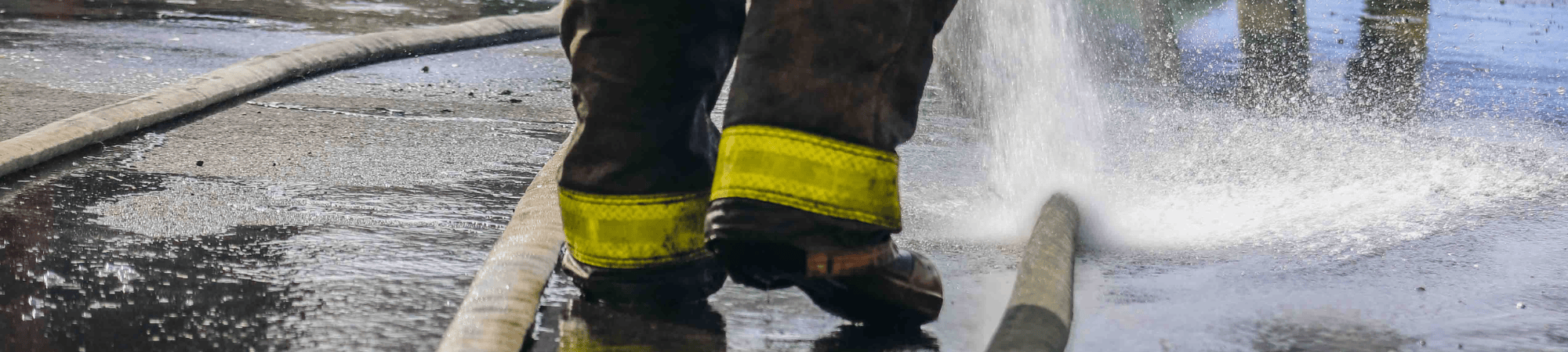 A firefighter's boots can be seen as water sprays on pavement with fire hoses lying on it.